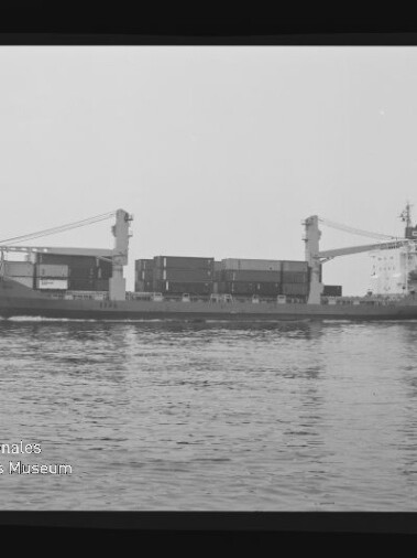 Schwarz-weiß-Seitenansicht eines Containerfrachtschiffs mit zwei Ladekränen und gestapelten Containern, das ruhig auf dem Meer liegt; Wasserzeichen des Internationalen Maritimen Museums Hamburg unten links.