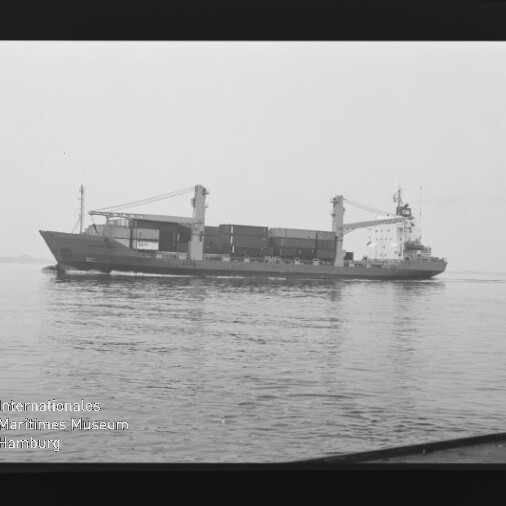 Schwarzweißaufnahme eines seitenansichtigen Containerschiffs mit zwei Ladekränen und gestapelten Containern auf ruhigem Wasser; Filmrand und Museumslogo unten links sichtbar.