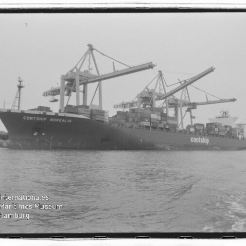 Schwarzweißaufnahme eines großen Containerschiffs (Contship Borealis) am Kai, von mehreren Hafenkranen beladen, mit aufgetürmten Containern auf dem Deck und ruhigem Hafenwasser im Vordergrund; links unten Wasserzeichen des Internationalen Maritimen Museums Hamburg.