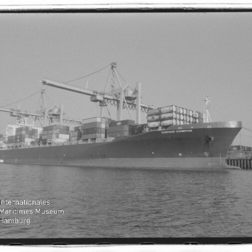 Schwarzweiß-Foto eines großen Containerschiffs am Kai mit übereinander gestapelten Containern und mehreren Hafenkränen, ruhigem Wasser im Vordergrund und einem Wasserzeichen des Internationalen Maritimen Museums Hamburg unten links.