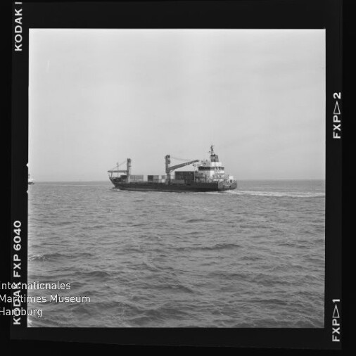 Schwarzweiß-Filmbild mit sichtbarem Kodak-Filmrand und Museumswasserzeichen, das ein mittleres Frachtschiff mit Ladekränen auf ruhiger See vor flachem Horizont zeigt.