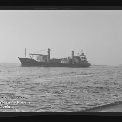 Schwarzweißaufnahme eines Containerschiffs mit aufgestapelten Containern und Ladekränen, das bei ruhiger See am Horizont fährt; im Vordergrund welliges Wasser, links unten Museumslogo und sichtbare Filmränder.