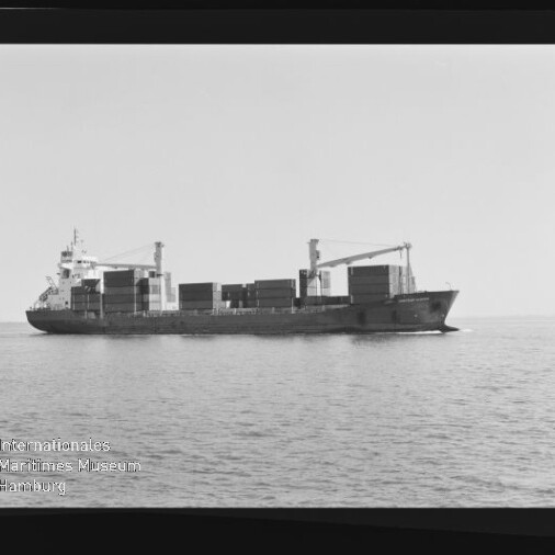 Schwarzweißfoto eines Containerschiffs in Seitenansicht auf ruhiger See mit aufgestapelten Containern und zwei Ladekränen an Deck, im Hintergrund ein kleines Schiff und unten links das Wasserzeichen ‚Internationales Maritimes Museum Hamburg‘.