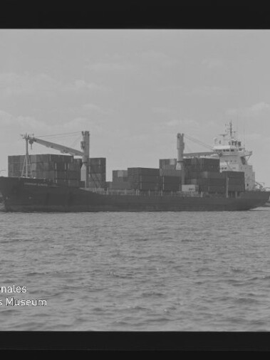 Schwarzweißaufnahme eines mit gestapelten Containern beladenen Frachtschiffs auf offenem Wasser, links ein Schlepper, im Hintergrund ein Pier oder eine Brücke und zwei Ladekräne, links unten das Logo des Internationalen Maritimen Museums Hamburg.
