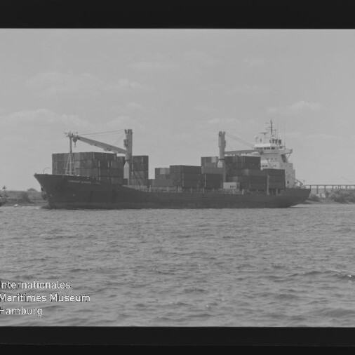 Schwarzweißaufnahme eines mit gestapelten Containern beladenen Frachtschiffs auf offenem Wasser, links ein Schlepper, im Hintergrund ein Pier oder eine Brücke und zwei Ladekräne, links unten das Logo des Internationalen Maritimen Museums Hamburg.