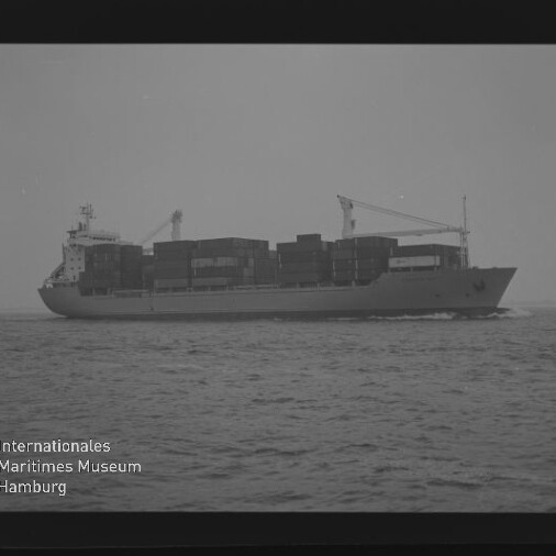 Schwarzweiß-Aufnahme eines Containerschiffs in Seitenansicht auf ruhiger See, mit gestapelten Containerblöcken und zwei Ladekränen auf dem Deck sowie sichtbarem Filmrand und Wasserzeichen des Internationalen Maritimen Museums Hamburg unten links.