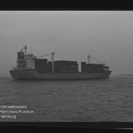 Schwarz‑weiß‑Foto eines beladenen Containerschiffs in Seitenansicht mit gestapelten Containern und zwei Ladekränen, das auf ruhiger See unter bedecktem Himmel fährt; am Bildrand Filmrand mit AGFA APX 100 und Museumswasserzeichen sichtbar.