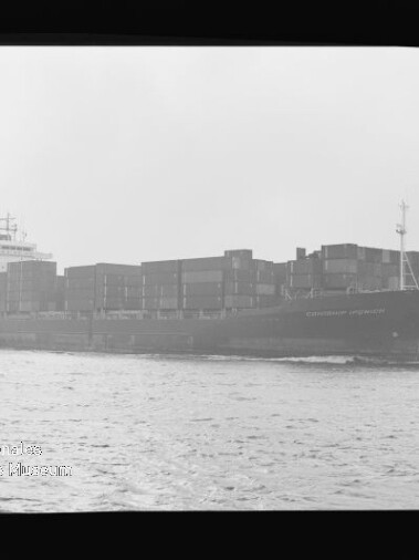 Schwarzweiß-Foto eines großen Containerschiffs auf offener See in seitlicher Ansicht, beladen mit gestapelten Containern; links unten ist ein Museumswasserzeichen sichtbar.