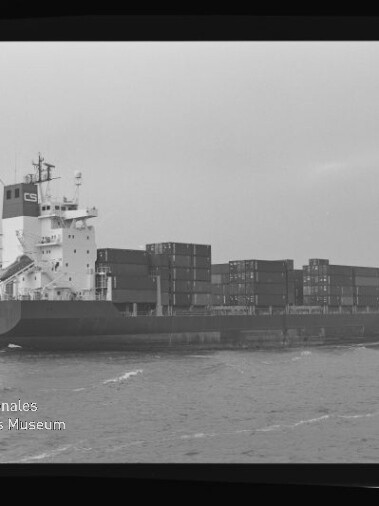 Schwarzweiß-Foto eines beladenen Containerschiffs auf See, seitlich aufgenommen mit übereinander gestapelten Containern, innerhalb eines Filmnegativ-Rahmens mit der rechten Beschriftung AGFA APX 100 und einem Wasserzeichen des Internationalen Maritimen Museums Hamburg unten links.