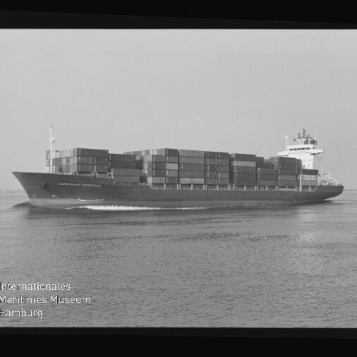 Schwarzweiß-Aufnahme eines großen Containerschiffs, beladen mit gestapelten Containerreihen, das bei ruhiger See mit sichtbarer Bugwelle dahinfährt; schwarzer Filmrand und Museumslogo links unten erkennbar.