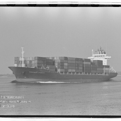 Schwarzweiß-Aufnahme eines beladenen Containerschiffs, das mit hohen Stapeln von Frachtcontainern auf ruhiger See fährt; am unteren linken Bildrand ein Wasserzeichen des Internationalen Maritimen Museums Hamburg.