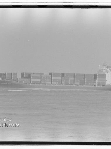 Schwarzweiß-Aufnahme eines Containerschiffs in Seitenansicht mit mehreren übereinander gestapelten Containerreihen, das ruhig auf dem Meer fährt; unten links sichtbar das Wasserzeichen des Internationalen Maritimen Museums Hamburg und ein Filmrandrahmen.