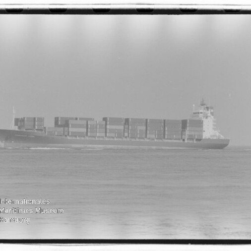 Schwarzweiß-Seitenansicht eines beladenen Containerschiffs auf ruhiger See mit gestapelten Containern und sichtbarer Bugwelle; unten links Wasserzeichen „Internationales Maritimes Museum Hamburg“.