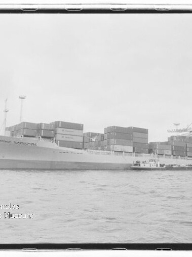 Großes Containerschiff mit gestapelten Ladecontainern liegt im Hafen, rechts Kräne im Hintergrund und ein kleines Begleitschiff längsseits, Wasser im Vordergrund und ein Wasserzeichen des Internationalen Maritimen Museums Hamburg unten links.