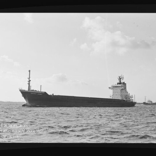 Großes Frachtschiff seitlich auf offener See mit achtern liegender Brücke und Mast am Bug, ruhige, leicht wellige See, bewölkter Himmel und ein weiteres Schiff am Horizont; Wasserzeichen des Internationalen Maritimen Museums Hamburg unten links.