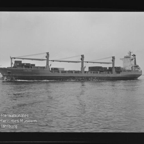Schwarzweiß-Foto einer Seitenansicht eines Frachtschiffs mit drei großen Ladekränen und auf dem Deck gestapelten Containern, das ruhig über dem Wasser fährt; am unteren linken Rand das Logo des Internationalen Maritimen Museums Hamburg und Rand des Negativfilms sichtbar.