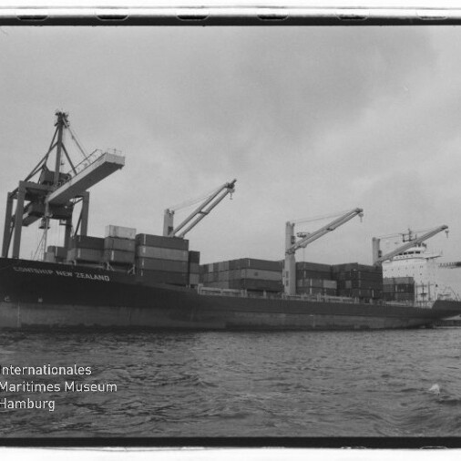 Großes Frachtschiff beladen mit Containern liegt im Hafen neben mehreren Ladekränen, Vordergrund Wasser und links unten sichtbares Logo des Internationalen Maritimen Museums Hamburg.
