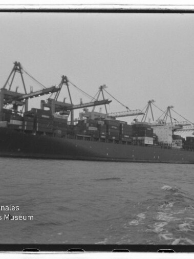 Schwarzweißfoto eines großen Containerschiffs am Kai, beladen mit gestapelten Containern und umgeben von mehreren Hafenkränen, aufgenommen vom Wasser aus; links unten das Signet des Internationalen Maritimen Museums Hamburg.