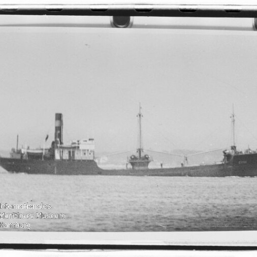 Schwarzweißaufnahme eines Frachtschiffs mit mittigem Schornstein und zwei Masten mit Ladebäumen auf ruhiger See, Wasserzeichen des Internationalen Maritimen Museums Hamburg unten links.