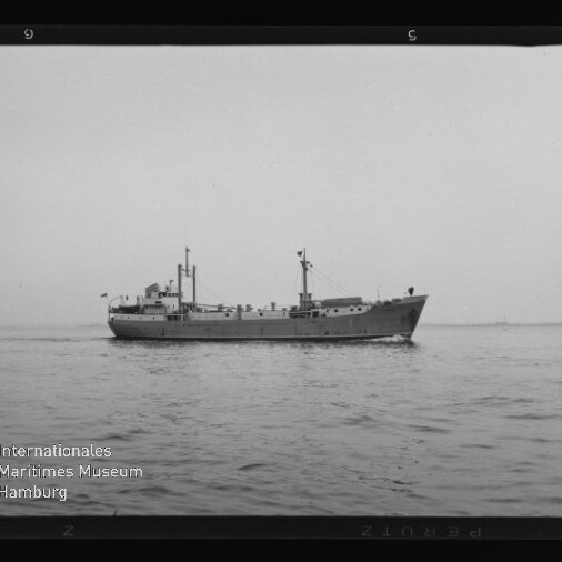 Schwarzweiß-Foto eines Frachtschiffs in Seitenansicht, ruhig auf dem Meer fahrend vor flachem Horizont mit sichtbaren Aufbauten, Masten und einer kleinen Flagge; im unteren linken Bildbereich das Wasserzeichen «Internationales Maritimes Museum Hamburg».