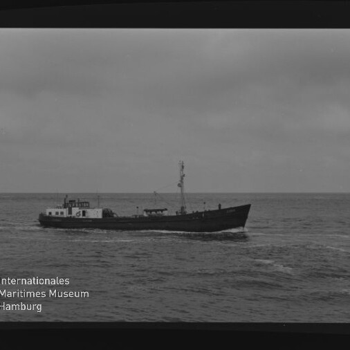 Schwarzweiß-Foto eines kleinen einmastigen Frachtschiffs, das auf ruhiger See unter bedecktem Himmel fährt; sichtbarer Filmrand mit Beschriftung AGFA PAN 100 rechts und Wasserzeichen des Internationalen Maritimen Museums Hamburg links unten.