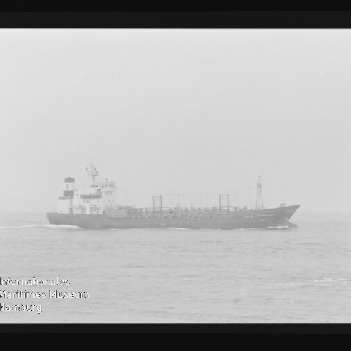 Großes Frachtschiff fährt bei diesigem, nebeligen Wetter auf ruhiger See, mit sichtbarer Bugwelle; Foto zeigt außerdem Filmrandmarkierungen und ein Wasserzeichen des Internationalen Maritimen Museums Hamburg.