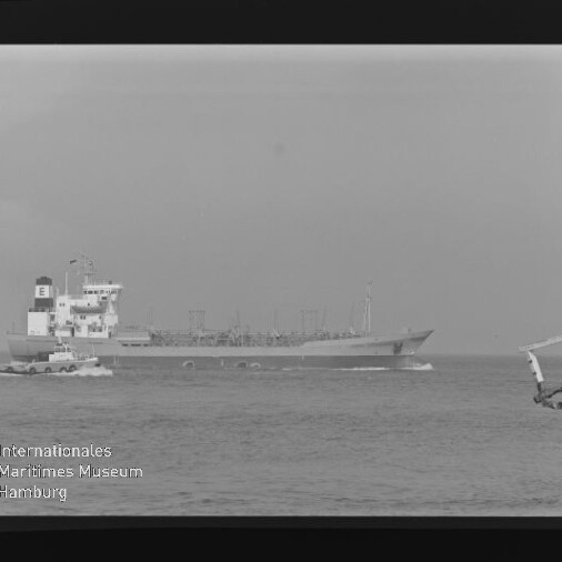 Schwarzweißaufnahme eines großen Tankschiffs mit Aufbauten am Heck, das auf ruhiger See entlangfährt und von einem kleinen Lotsenboot begleitet wird; rechts im Bild ein Pierfragment und unten links das Logo des Internationalen Maritimen Museums Hamburg.