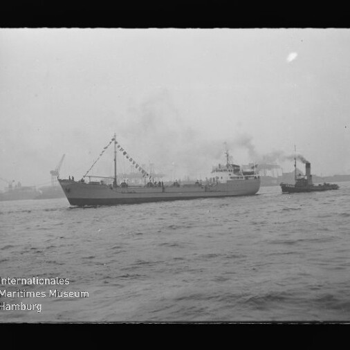 Schwarzweißaufnahme eines beflaggeten Frachtschiffs, das von einem Schlepper durch einen nebligen Industriehafen mit rauchenden Schornsteinen und Kränen begleitet wird; unten links das Logo des Internationalen Maritimen Museums Hamburg.