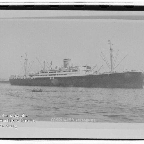 Schwarzweißfoto eines großen Passagier-/Frachtschiffs Cordillera (Hamburg) auf ruhigem Wasser, sichtbar sind Schornstein, Ladekräne und ein kleines Ruderboot im Vordergrund sowie ein Museumswasserzeichen in der linken unteren Ecke.