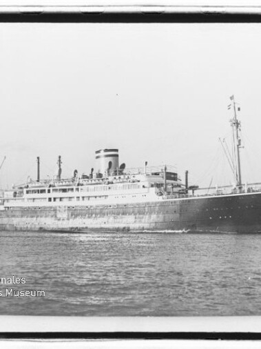 Schwarzweißfoto eines großen historischen Passagierschiffs mit einem einzelnen Schornstein und mehreren Lade- bzw. Masten, das auf ruhiger See entlangfährt; unten links sichtbares Museumssignet.