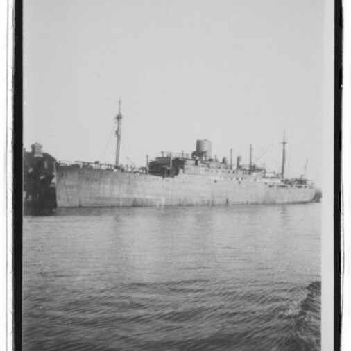 Schwarzweiß-Foto eines großen grauen Frachtschiffs mit Schornstein und mehreren Masten, längsseits an einem Kai bei ruhigem Wasser; unten links das Logo des Internationalen Maritimen Museums Hamburg.