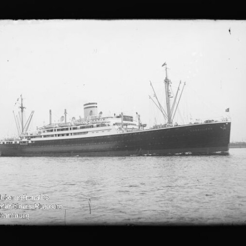 Schwarzweiß-Foto eines großen Passagierdampfers im Seitenprofil auf ruhiger See vor einem Hafen, mit hohem Schornstein, mehreren Ladebäumen und einer Flagge am Heck; unten links ein runder Museumsstempel und ein schwarzer Negativrahmen um das Bild.