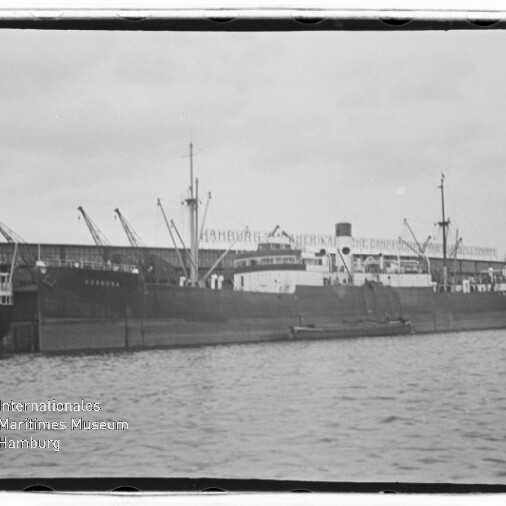Schwarz‑weiß-Foto eines großen Frachtschiffs mit Masten und Schornstein, vertäut am Kai vor einem Lagerhaus mit mehreren Ladekränen, Wasser im Vordergrund und einem 'Internationales Maritimes Museum Hamburg'-Wasserzeichen unten links.