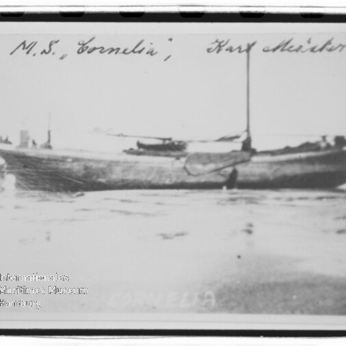 Schwarzweiß-Fotografie eines hölzernen Segelboots namens „Cornelia“, seitlich in seichtem Wasser oder am Strand liegend, mit aufragendem Mast, handschriftlicher Beschriftung am oberen Rand und Museumswasserzeichen unten links.