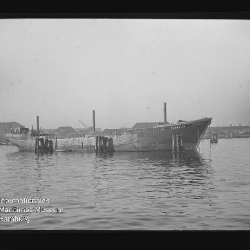 Schwarzweiß-Foto eines länglichen Frachtschiffs, das ruhig zwischen hölzernen Dalben in einem Industriehafen liegt, vor niedrigen Lagergebäuden im Hintergrund und mit dem Schriftzug des Internationalen Maritimen Museums Hamburg in der unteren linken Ecke.