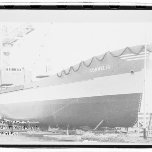 Großes Frachtschiff mit dem Namen CORNELIA im Trockendock, Bug mit Girlanden geschmückt, sichtbarer Rumpf und im Hintergrund Werftkran und Gerüste.
