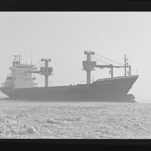 Schwarzweiß-Fotografie eines Frachtschiffs mit mehreren Deckskranen, das als dunkle Silhouette durch eisbedecktes Wasser fährt; am Bildrand sind Filmrandbeschriftungen (AGFAPAN 100) sichtbar.