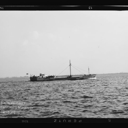 Schwarzweißaufnahme eines flachen Frachtschiffs mit mittigem Mast und Ladebaum, das auf ruhiger See nahe dem Horizont fährt; im Vordergrund wellige Wasseroberfläche und unten links ein Wasserzeichen des Internationalen Maritimen Museums Hamburg.