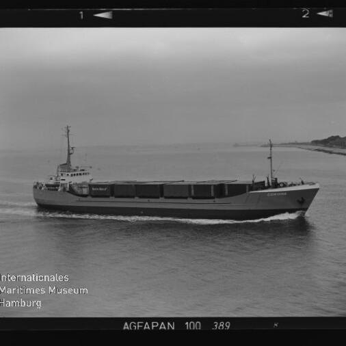 Schwarzweißfotografie eines Frachtschiffs mit mehreren Laderäumen und kleinem Mast, das nahe der Küste über ruhiges Wasser fährt und eine Heckwelle zieht; Bild im Filmrahmen mit Beschriftung AGFAPAN 100 und Wasserzeichen des Internationalen Maritimen Museums Hamburg.