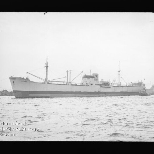 Schwarzweißaufnahme eines großen Frachtschiffs seitlich im Hafen, mit Ladekränen und Aufbauten, Wasser im Vordergrund und kleinem Schlepper am Heck; Wasserzeichen des Internationalen Maritimen Museums Hamburg links unten.