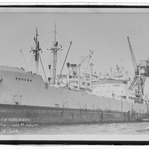 Schwarzweißfoto des Frachtschiffs CORONA, längsseits an einem Industriepier mit mehreren Ladekränen und Verladearmen, mit einem Schlepper am Heck und dem Logo des Internationalen Maritimen Museums Hamburg unten links.