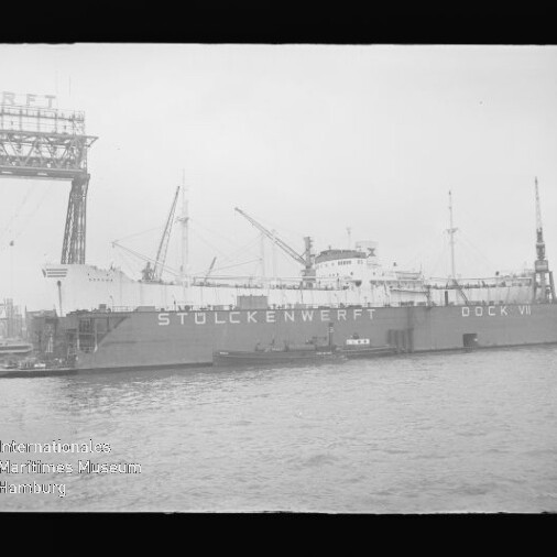 Großes Frachtschiff in einem Dock, an der Dockwand die Aufschrift STÜLCKENWERFT DOCK VI, mit mehreren Kränen und Hafenanlagen im Hintergrund und Wasser im Vordergrund.