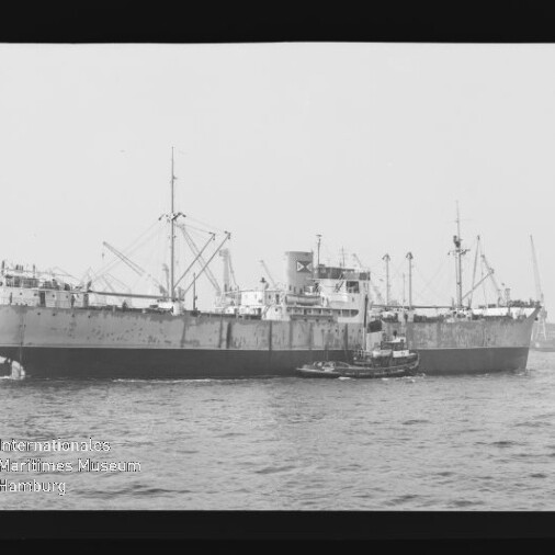 Schwarz‑weiß-Foto eines großen Frachtschiffs mit mehreren Masten und Ladekränen, begleitet von einem kleinen Schlepper auf dem Wasser; Filmrand und ein Wasserzeichen des Internationalen Maritimen Museums Hamburg links unten sind sichtbar.