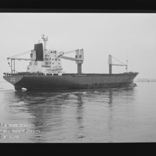 Schwarzweißaufnahme eines Frachtschiffs mit zwei auffälligen Ladekränen und aufragenden Aufbauten in der Bildmitte, das ruhig über spiegelndes Wasser fährt; unten links das Logo und der Schriftzug Internationales Maritimes Museum Hamburg.