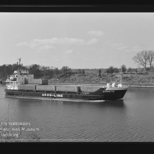 Schwarzweißaufnahme eines Containerschiffs namens CORVETTE mit der Aufschrift AROS-LINE am Rumpf, das mit Containern beladen einen Kanal entlangfährt; im Hintergrund Ufer mit Bäumen und unten links ein Museumswasserzeichen.