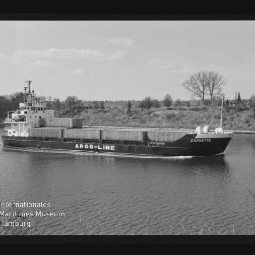 Schwarzweiß-Foto eines Containerschiffs namens „Corvette“ mit großer Aufschrift „AROS-LINE“, das ruhig einen Kanal passiert; im Hintergrund flaches Ufer mit Bäumen, links unten Museumssignet und sichtbarer Filmrahmenrand.