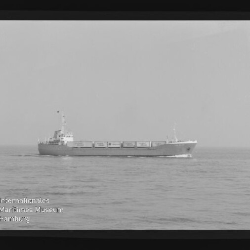 Schwarzweißaufnahme eines mittleren Frachtschiffs, das auf ruhiger See vor einem flachen Horizont entlangfährt; am linken unteren Bildrand ist das Logo des Internationalen Maritimen Museums Hamburg sichtbar.