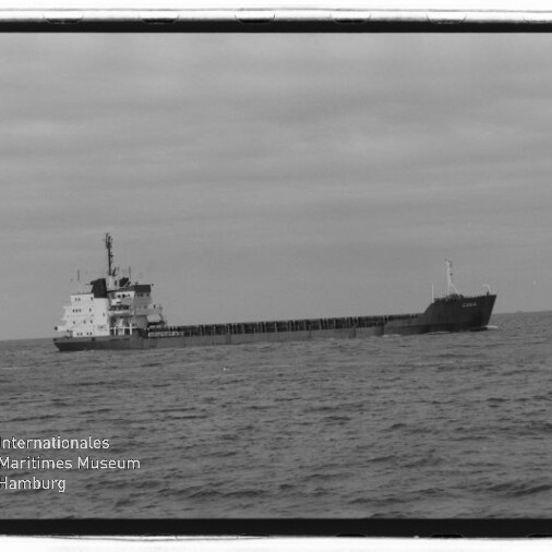 Schwarzweißaufnahme eines langen Frachtschiffs mit achtern liegenden Aufbauten, das bei ruhiger See am Horizont fährt; im linken unteren Eck das Wasserzeichen des Internationalen Maritimen Museums Hamburg.