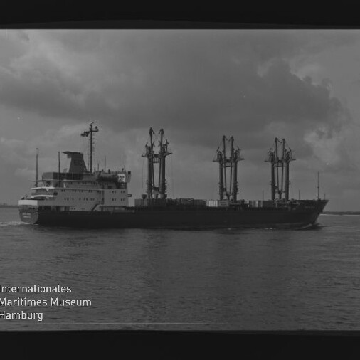 Schwarzweißaufnahme eines Frachtschiffs mit drei hohen Ladekränen und achterem Aufbauten, das auf ruhigem Wasser unter wolkenverhangenem Himmel fährt; Logo des Internationalen Maritimen Museums Hamburg unten links.