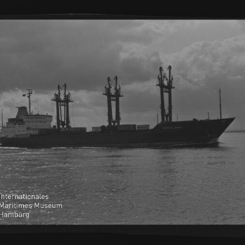 Schwarzweiß-Foto eines Frachtschiffs mit drei hohen Ladekränen, das auf ruhigem Wasser unter wolkigem Himmel fährt; zu sehen sind Filmrand und Wasserzeichen des Internationalen Maritimen Museums Hamburg.